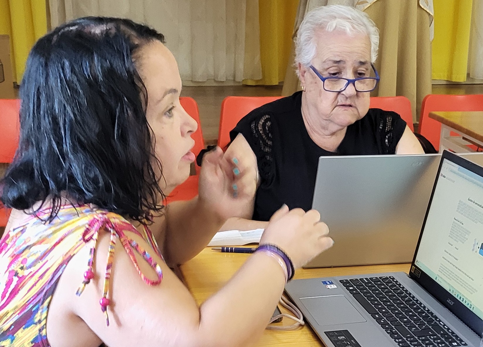 Dos mujeres usando portátiles en aula, una joven explicando a una mayor durante una sesión de aprendizaje digital